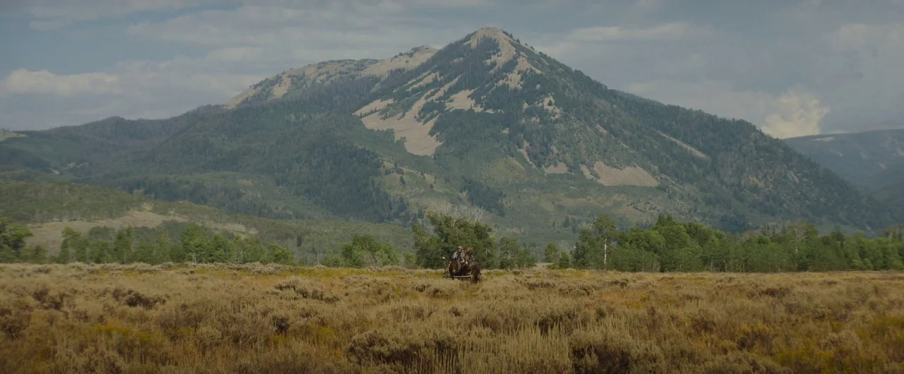 Damsel (2018) movie still: wide — A vast, dry sagebrush valley sits before a large, prominent mountain under cloudy skies. Two men sit on a wooden…