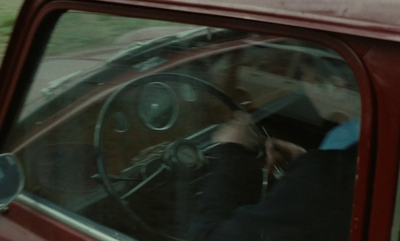 Graduate First (1978) movie still: medium — The interior of a vintage car with a wood-grain dashboard and thin steering wheel. A person in a dark…