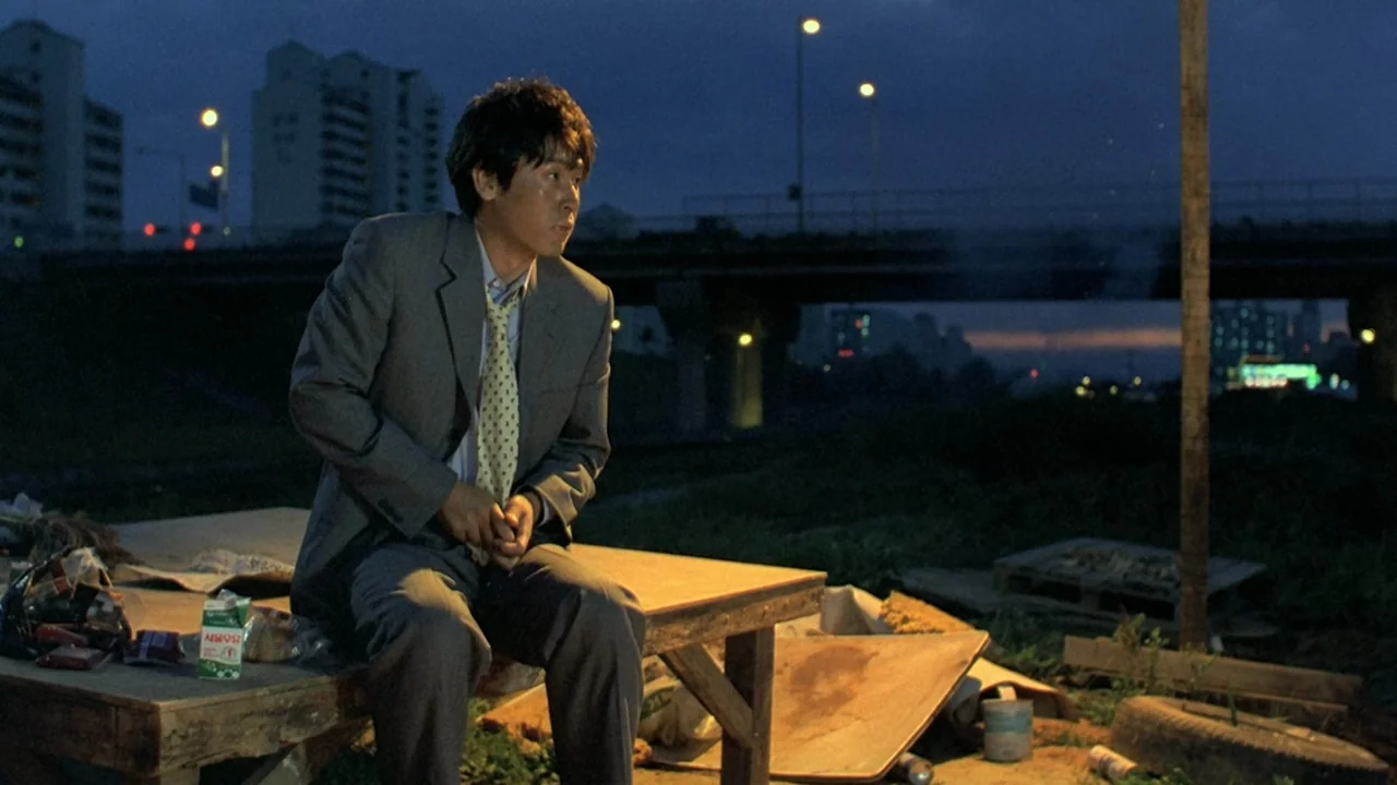 Peppermint Candy (1999) movie still: medium — A man sits on a wooden platform beside a river at night under city lights. An overpass looms in the…
