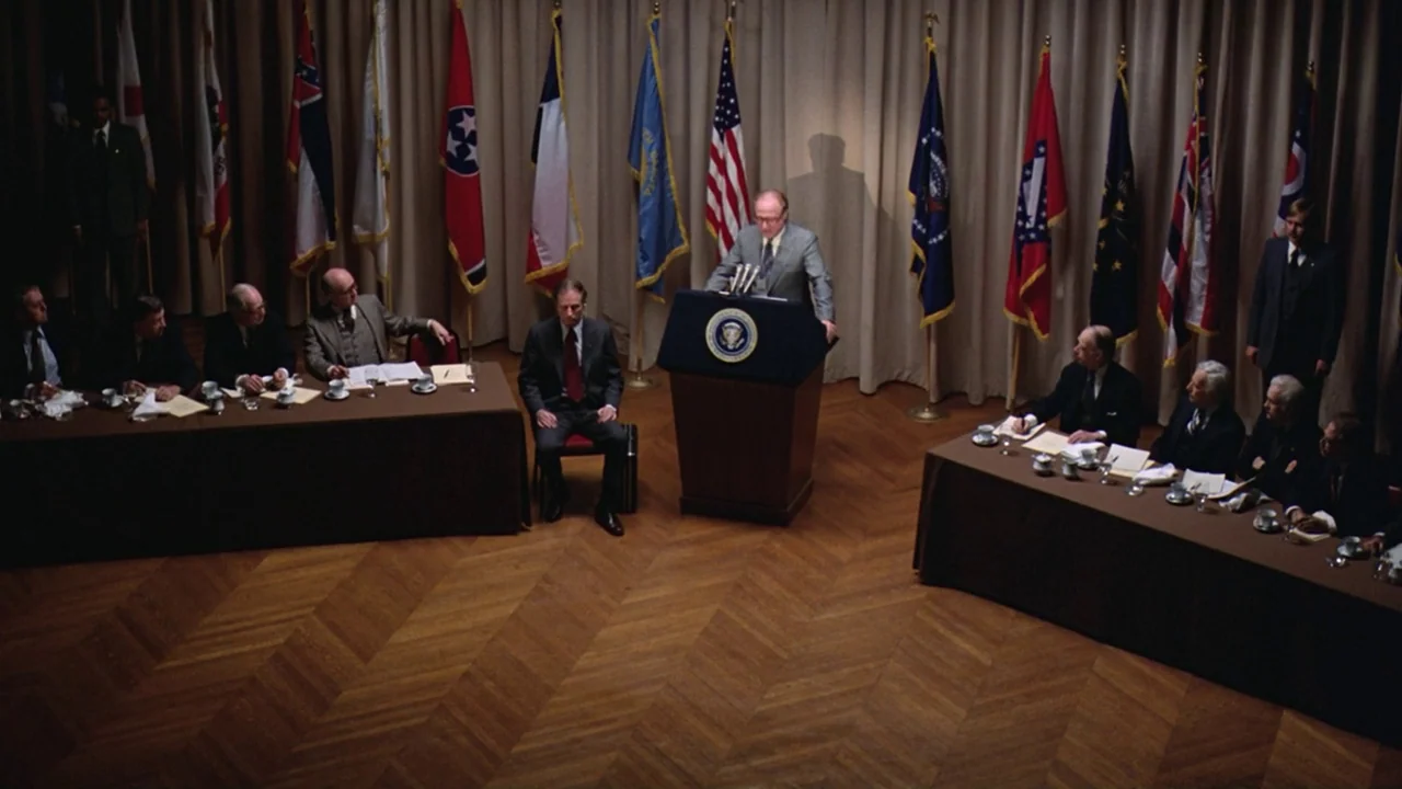 Being There (1979) movie still: wide — A formal room with parquet wooden floors is lined with a series of state flags. A man addresses a seated…