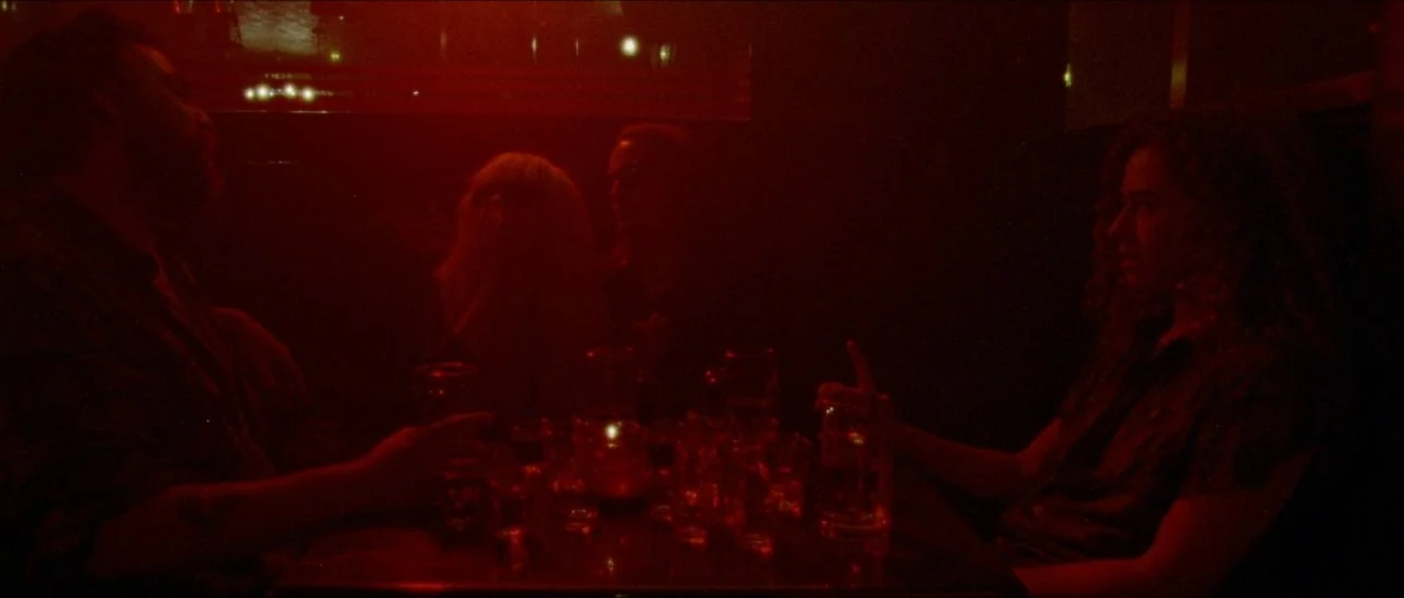 Bliss movie still: medium — A dimly lit bar booth filled with intense red light and glass surfaces. Two men sit across from each other at a small…