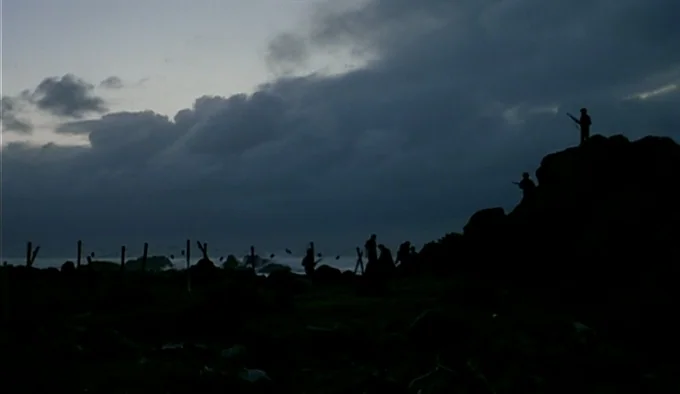 Dust In The Wind (1986) movie still: wide — A rocky coastal ridge sits under a dark, overcast twilight sky. Soldiers stand in silhouette against the…