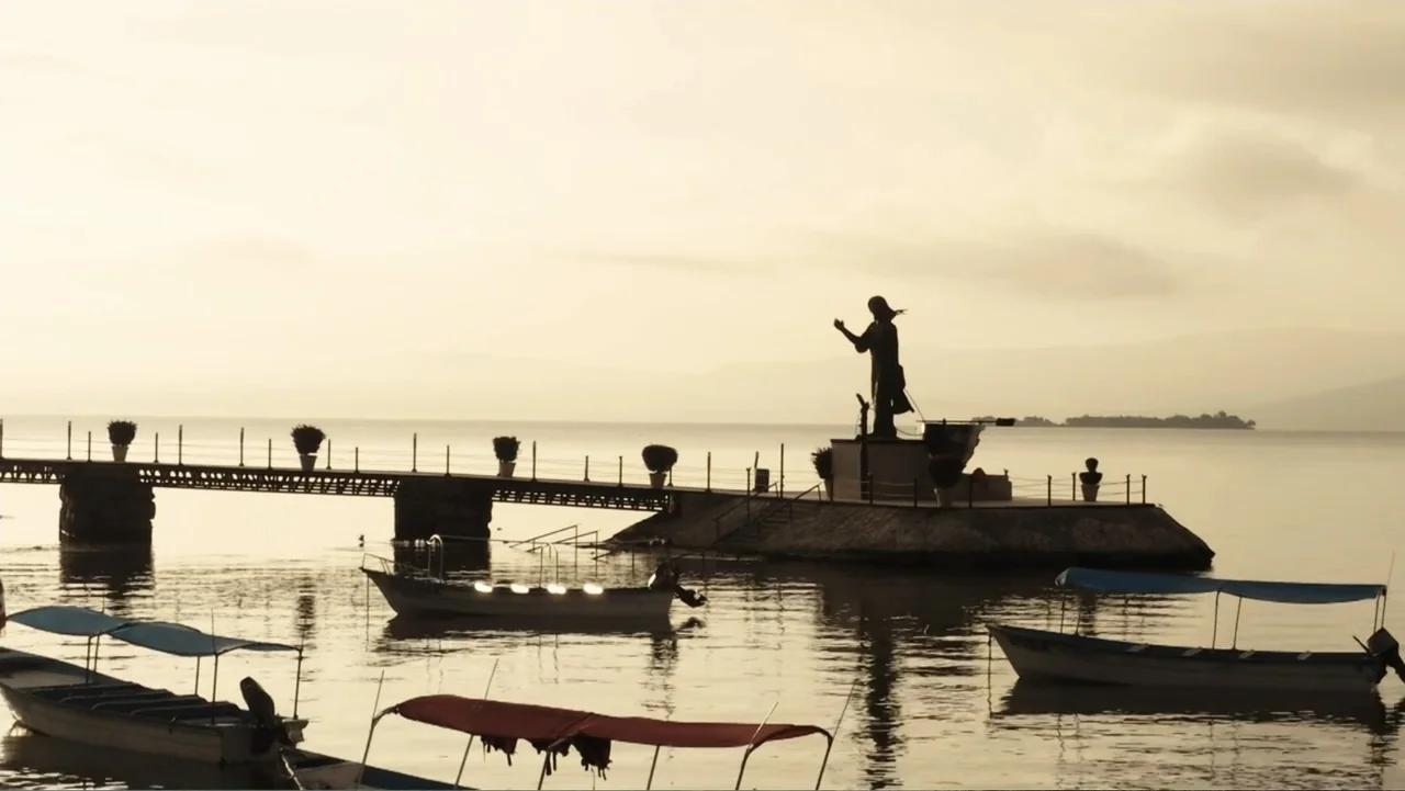 Por Amor al Money (2023) movie still: wide — A stone pier extends into a calm lake with a central statue at the end. Small wooden boats with canopy…