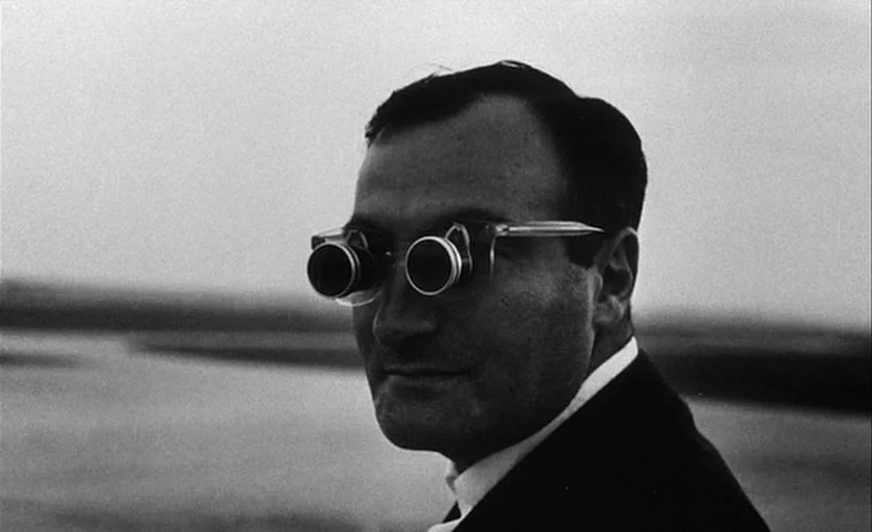 La Jetée (1962) movie still: closeup — An outdoor coastal area with a blurry shoreline in the background. A man wears dual-lens spectacles while…