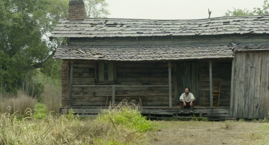 Free State of Jones (2016) movie still: wide — A weathered wooden log cabin features a damaged shingled roof and a front porch. A lone man sits…