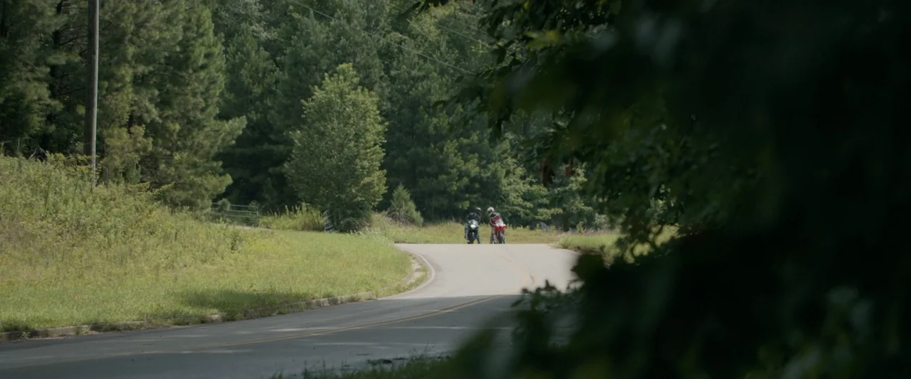 One Fast Move (2024) movie still: wide — A rural asphalt road curves through a dense forest with grassy shoulders and trees. Two motorcyclists ride…