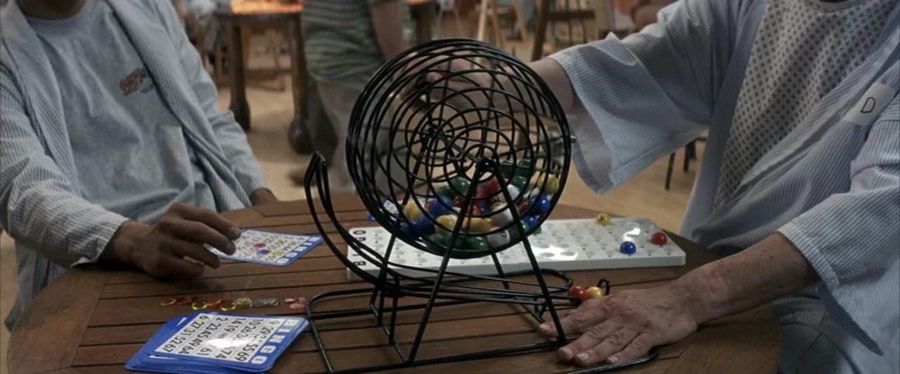 50 First Dates (2004) movie still: medium — Two individuals sit at a wooden table in a room with light wood floors. They are engaged in a bingo game…