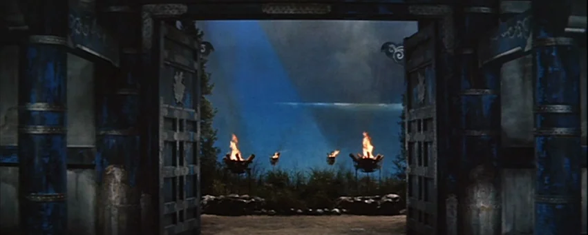 Kwaidan (1964) movie still: establishing — A stone temple portal framed by ornate blue columns opens to a night landscape. Four iron braziers burn…