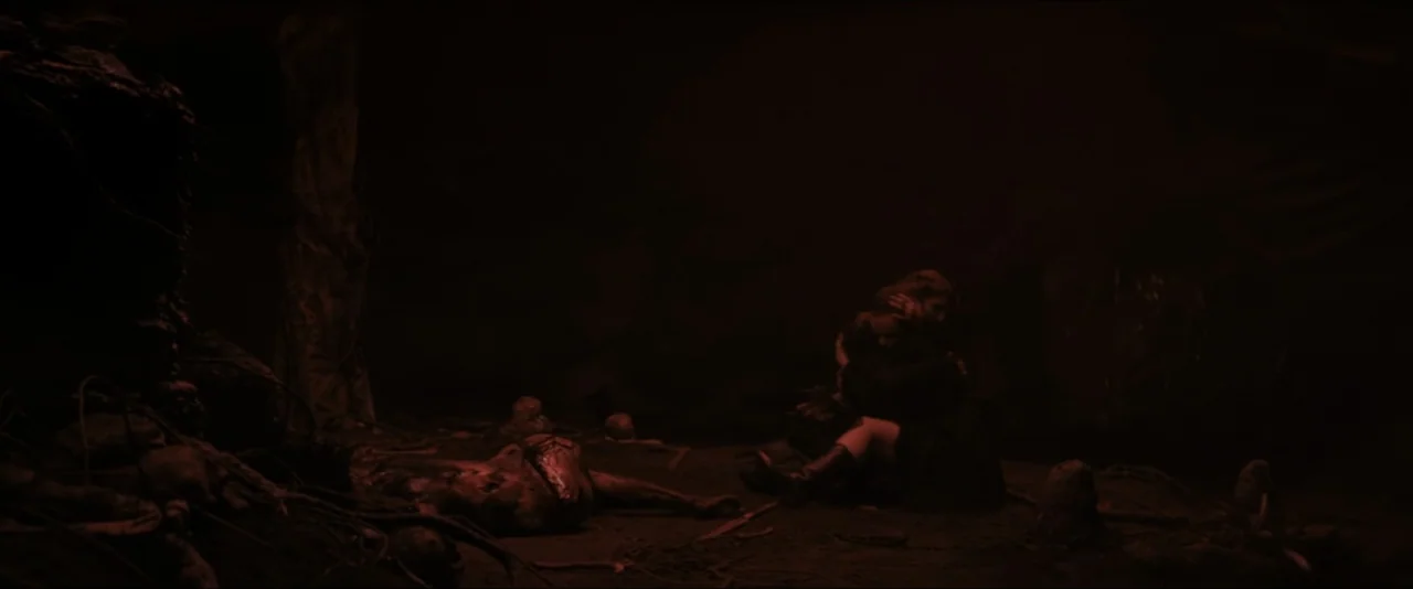 It Feeds (2025) movie still: wide — A dark, cavernous space is filled with shadows and indistinct debris on the ground. A woman sits on the floor…