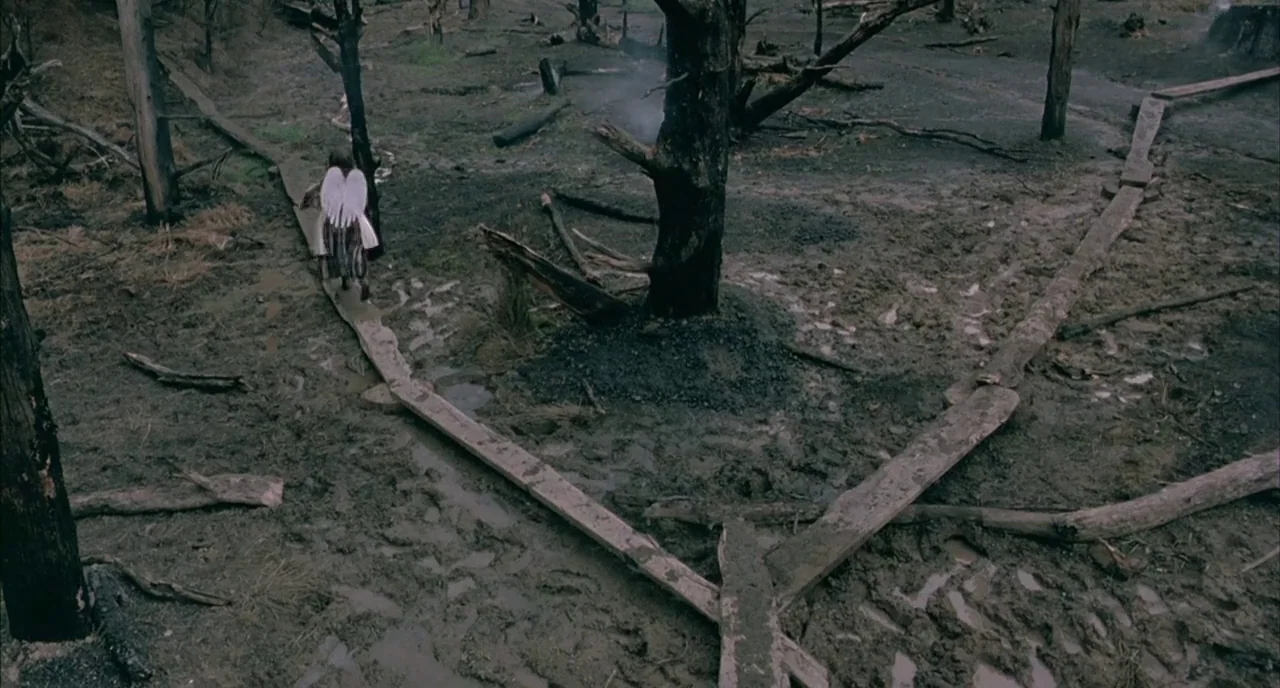 The Piano (1993) movie still: wide — A muddy, desolate terrain is scattered with scorched tree trunks and wooden planks. A small girl wearing white…
