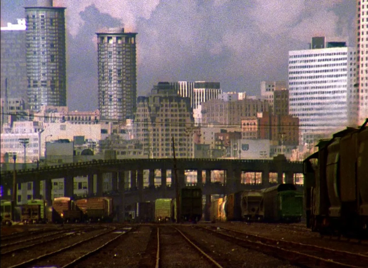 Streetwise (1984) movie still: establishing — An industrial railway yard stretches toward a dense urban skyline of varied office towers. Several…