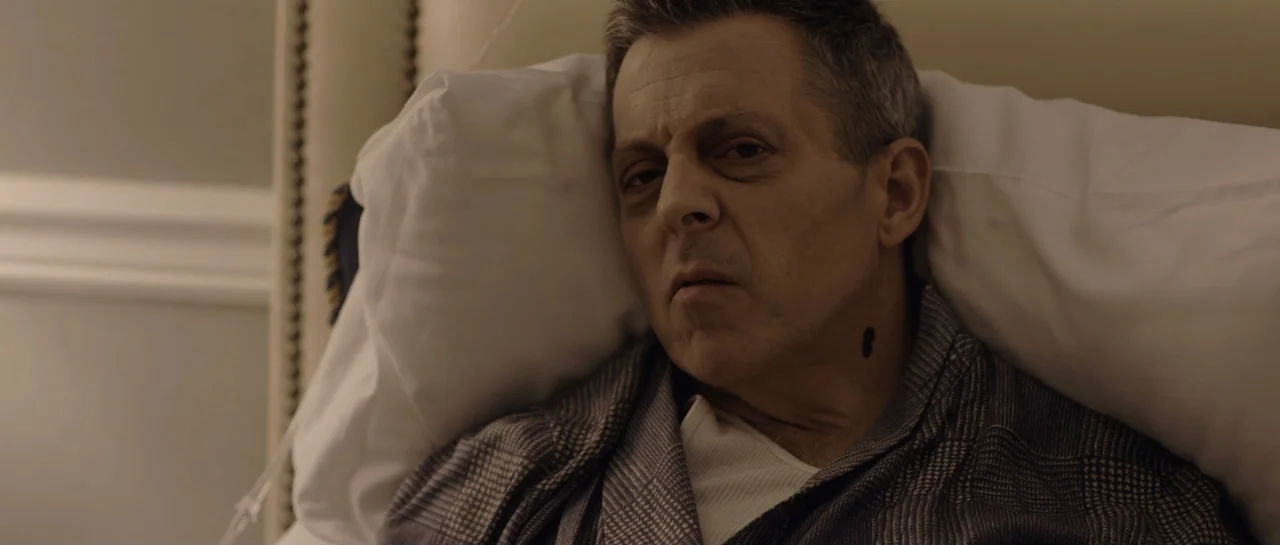 NY84 (2016) movie still: closeup — A sterile hospital room environment features white pillows and medical surroundings. A middle-aged man with short…