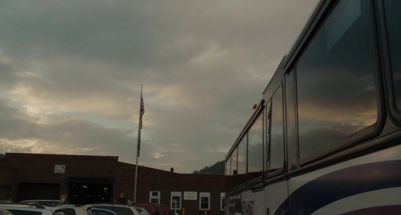 Paterson (2016) movie still: wide — A bus depot parking lot features a brick building under a cloudy evening sky. The side of a bus dominates the…