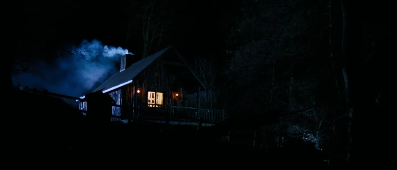 The Descent (2005) movie still: wide — A wooden cabin sits in a dark forest at night with smoke billowing from its chimney. Warm light shines from…