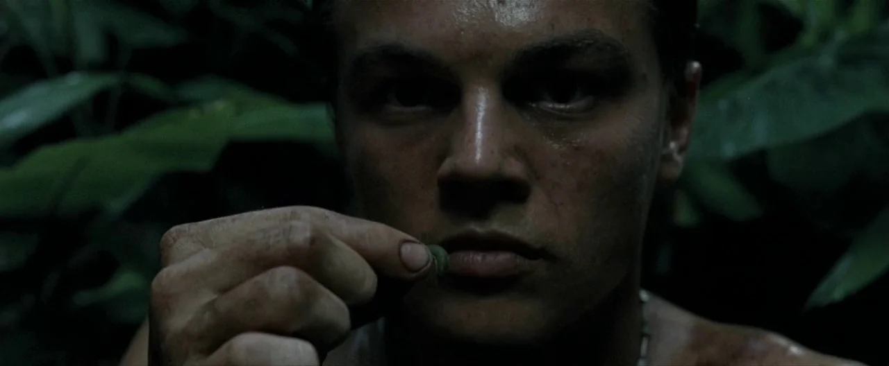 The Beach (2000) movie still: closeup — A dense, dark jungle environment filled with out-of-focus tropical leaves. A young man holds a small green…