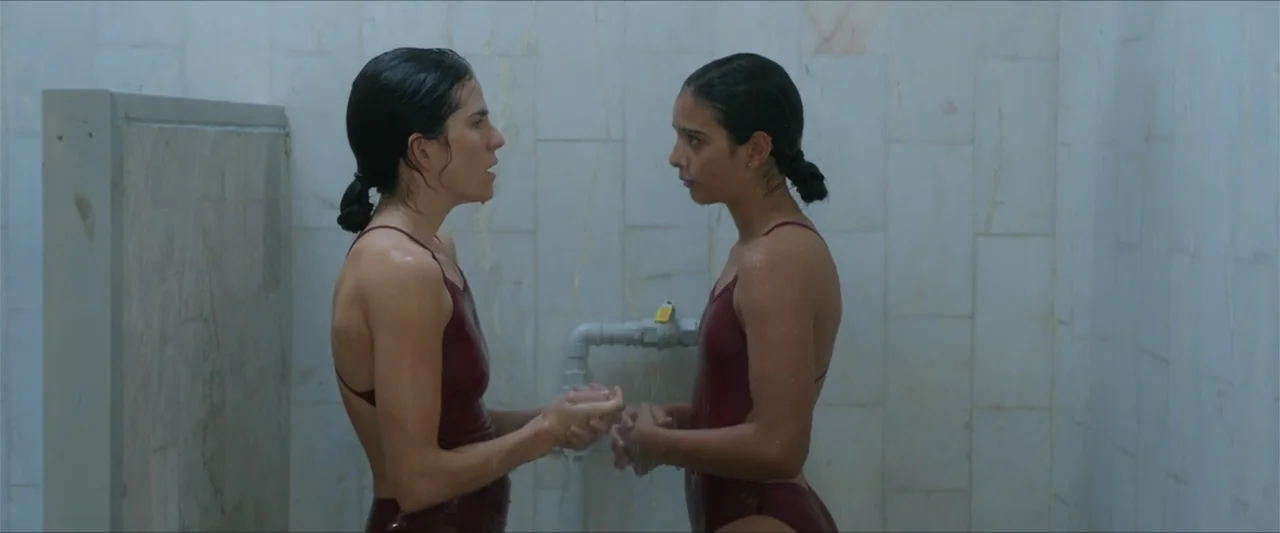 Dive (2013) movie still: medium — A public shower area features light-colored marble wall tiles and industrial white plumbing fixtures. Two women in…