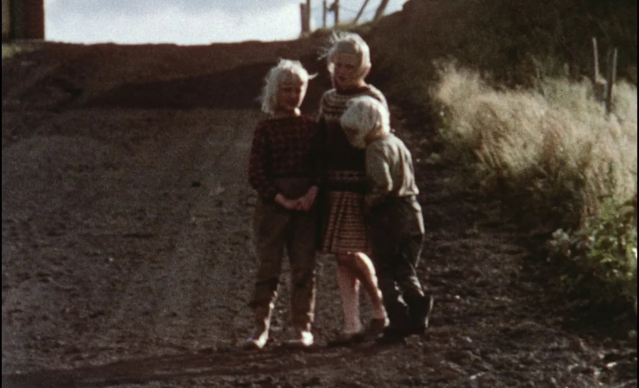 Sans Soleil (1983) movie still: fullbody — Three children stand clustered together in the center of an unpaved dirt path. Tall dry grass grows along…