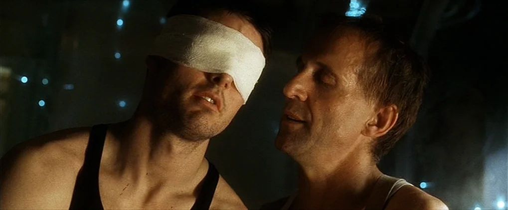 Minority Report movie still: closeup — A dimly lit interior features two men in tank tops standing close together. One man wears a bandage over his…