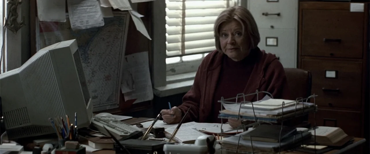 Angst (2017) movie still: medium — A cluttered 1990s office features a beige computer monitor, hanging papers, and filing cabinets. An elderly woman…