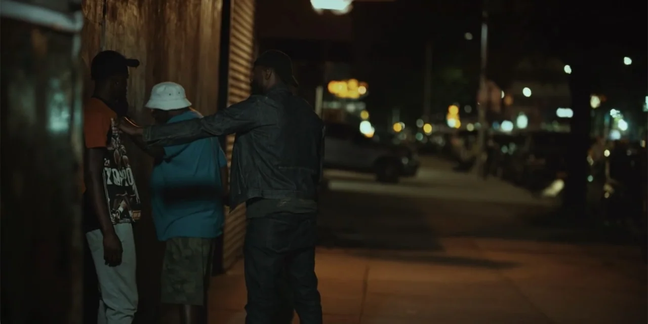 Equal Standard (2021) movie still: medium — Three men stand on a dimly lit urban sidewalk near a corrugated metal wall. One man gestures toward the…