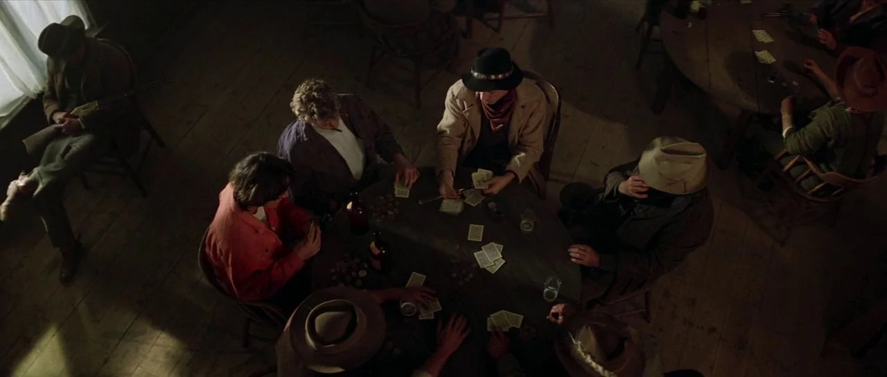 Bill and Ted’s Excellent Adventure (1989) movie still: wide — A dimly lit wooden saloon room contains multiple men seated at circular tables. The…