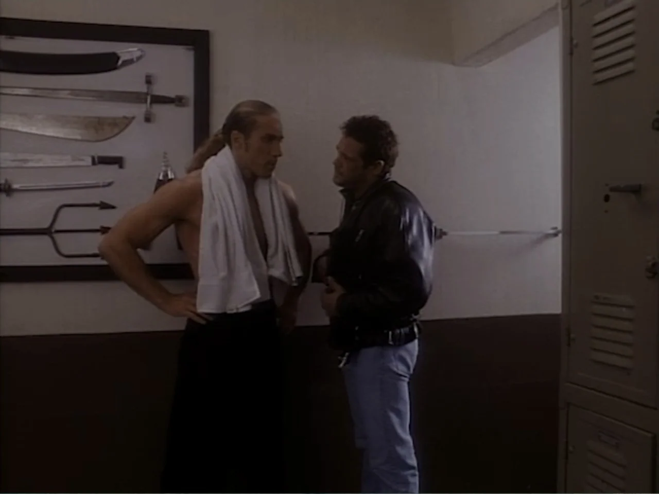 Firepower (1979) movie still: medium — A locker room contains a framed display of various swords and weapons mounted on wall. Two men stand facing…