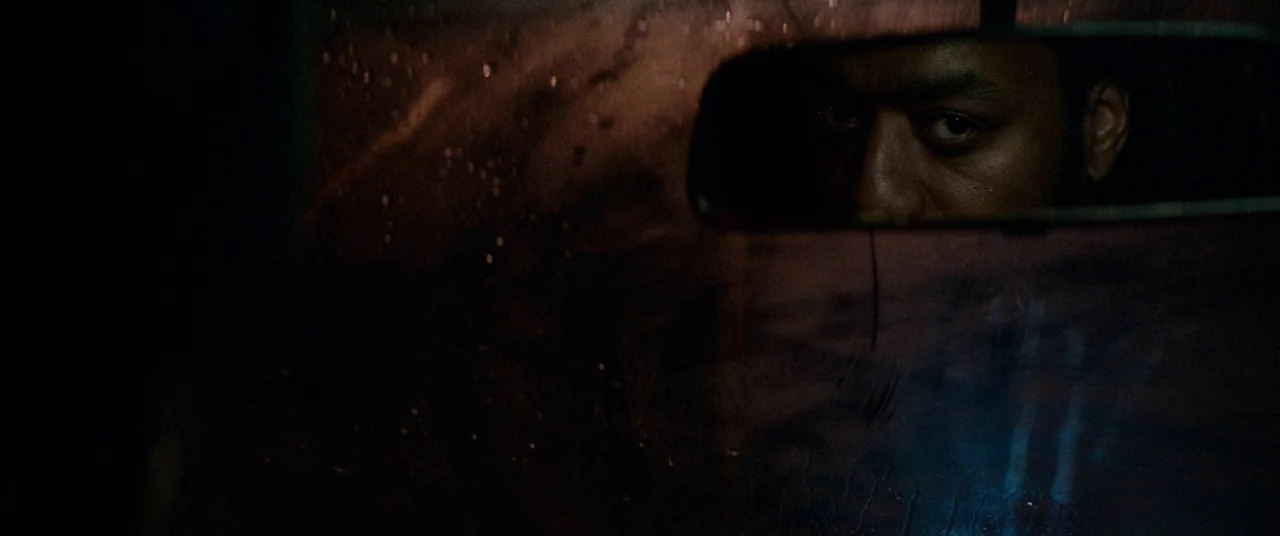 Triple 9 (2016) movie still: closeup — The interior of a vehicle at night featuring a rain-streaked windshield. A man is visible as a reflection in…