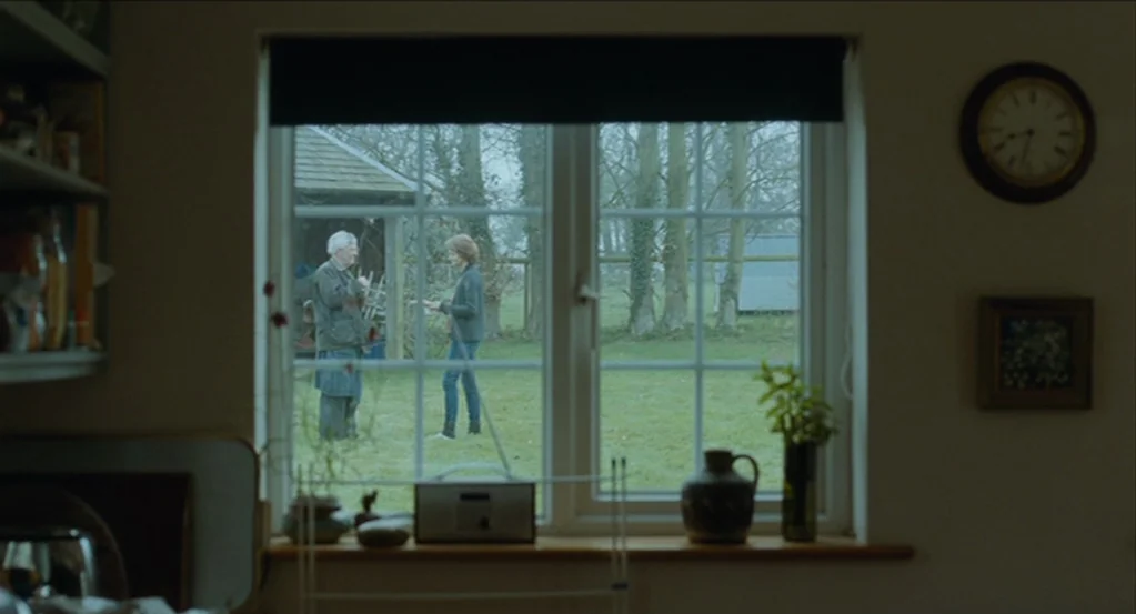 45 Years (2015) movie still: medium — A view through an interior window looks out into a grassy backyard area. Two older individuals stand on the…