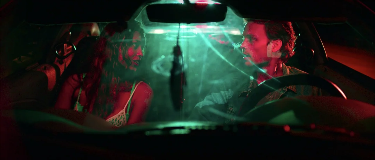 68 Kill (2017) movie still: medium — The interior of a parked car at night is bathed in dramatic colored lighting. A man and a woman face each other…