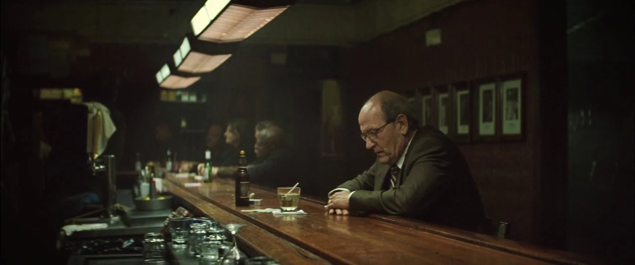 Killing Them Softly (2012) movie still: medium — A dimly lit bar features a long, polished wooden counter and overhead light fixtures. A balding man…