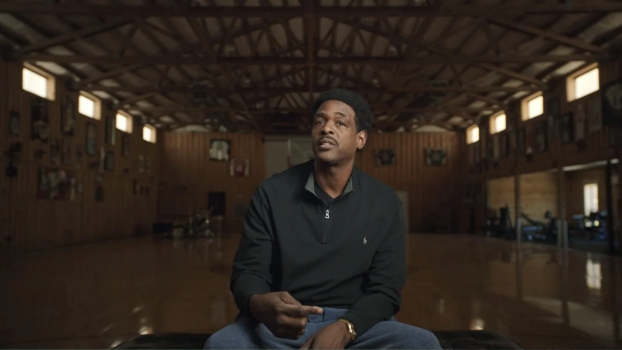 We Beat the Dream Team (2025) movie still: medium — A spacious wooden gymnasium with exposed beams and framed pictures along the walls. A man in a…