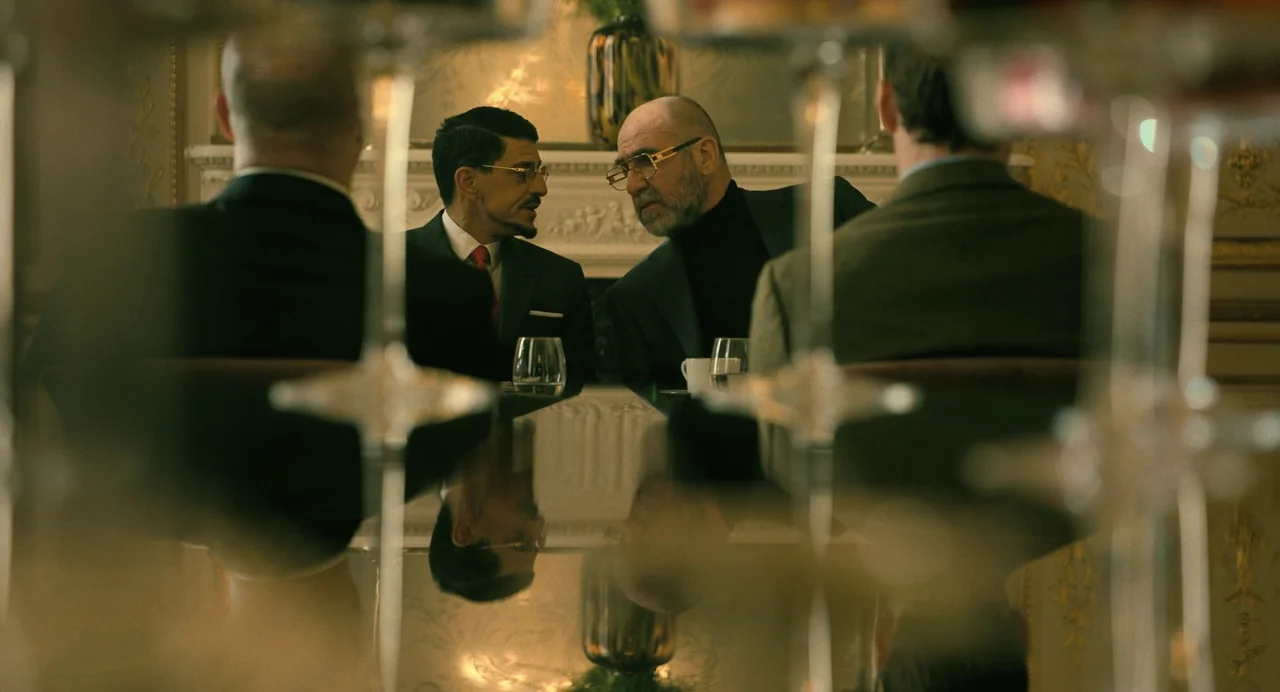 The Killer (2024) (2024) movie still: medium — An opulent room with gold-accented wallpaper features a dark reflective table. Two men sit across…