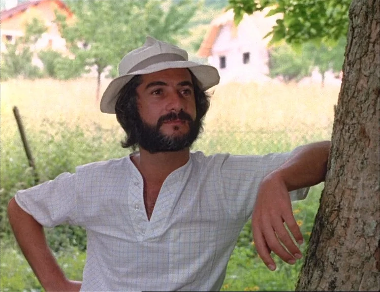Claire’s Knee (1970) movie still: medium — A man stands in a grassy field with rural dwellings visible in the background. The man leans against a…
