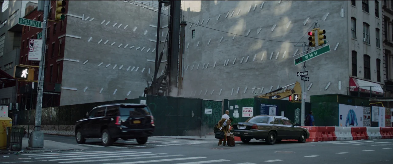 Tár (2022) movie still: wide — A busy urban intersection in New York featuring construction scaffolding and grey concrete walls. Two men are placing…