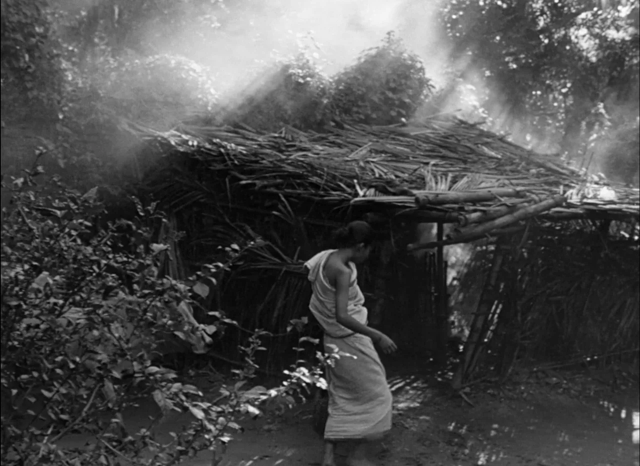 Pather Panchali (1955) movie still: medium — A rural setting features a small, deteriorating structure with a roof made of palm fronds. Smoke rises…