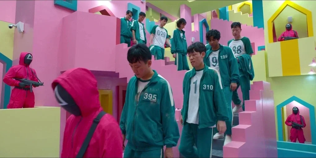 Making of: Squid Game (2021) movie still: wide — A geometric stairwell interior features pastel pink walls and vibrant blue and yellow architectural…