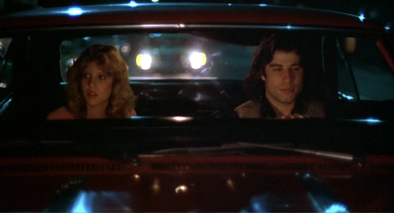 Carrie (1976) movie still: medium — A dark vehicle interior is illuminated by headlights shining through the rear windshield. A young man drives the…