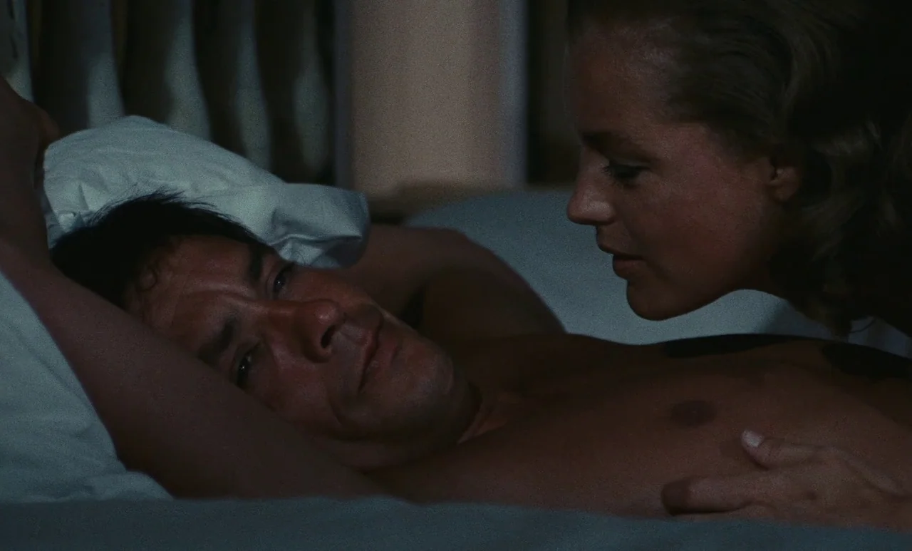 The Swimming Pool (1969) movie still: medium — A dimly lit setting shows two individuals resting in a bed with white pillows. The woman leans toward…