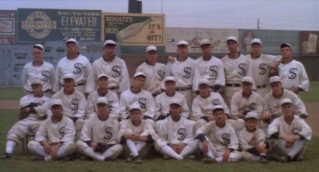Eight Men Out (1988) movie still: wide — A group of baseball players in vintage uniforms stands and sits in rows. Behind them is a scoreboard and…