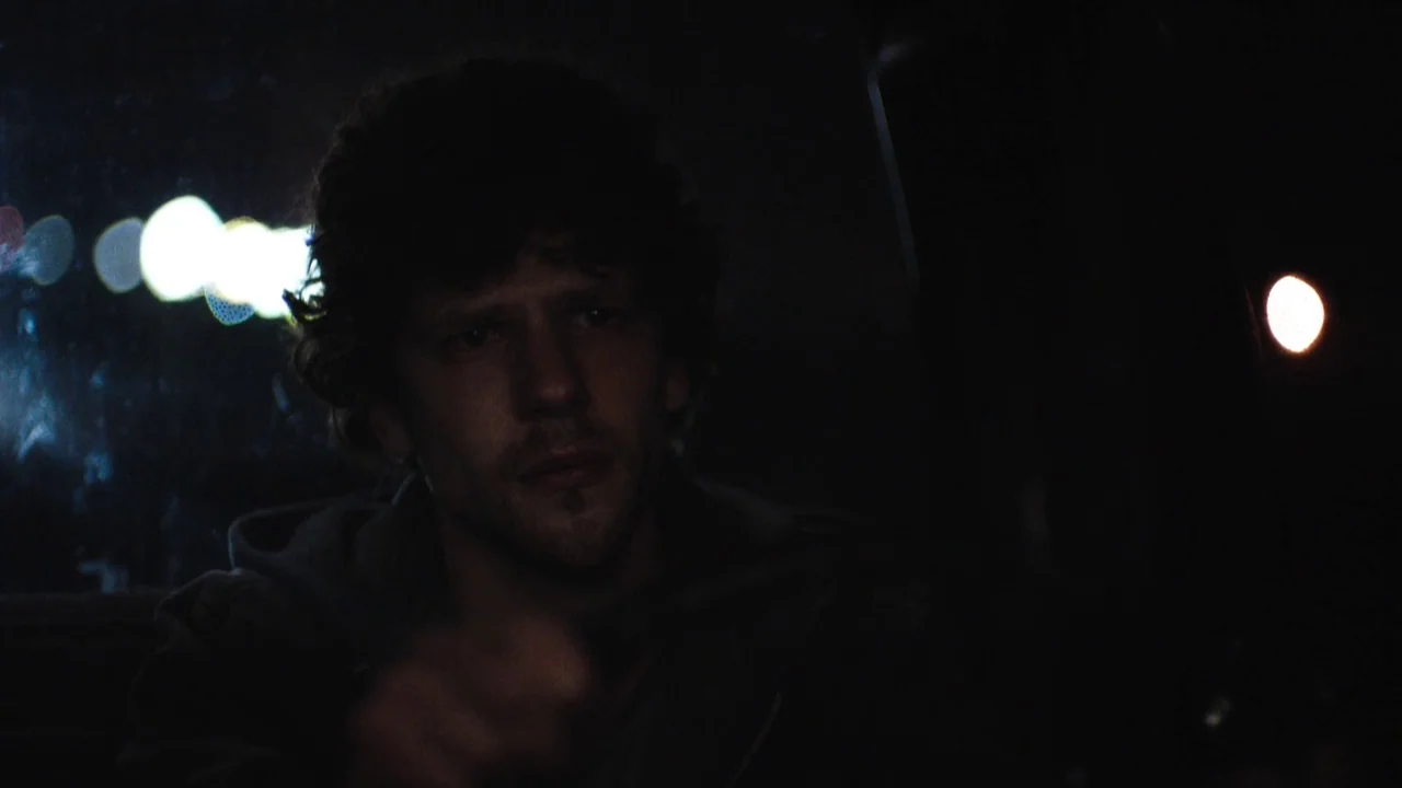 Night Moves (2013) movie still: medium — A dark car interior at night with blurred bokeh lights visible through the window. A young man with dark…