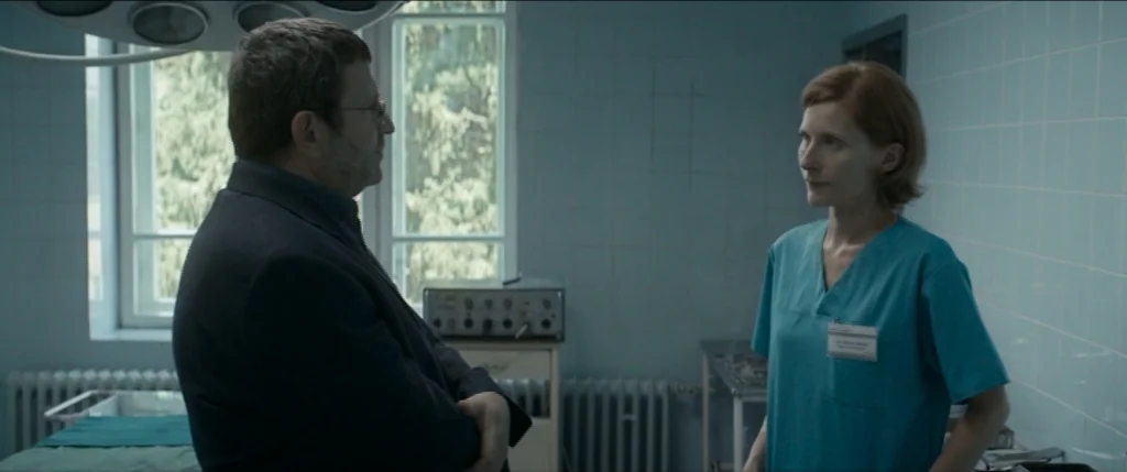 Graduation (2016) movie still: medium — An austere medical room with light blue tiled walls features medical equipment and windows. A man in a dark…
