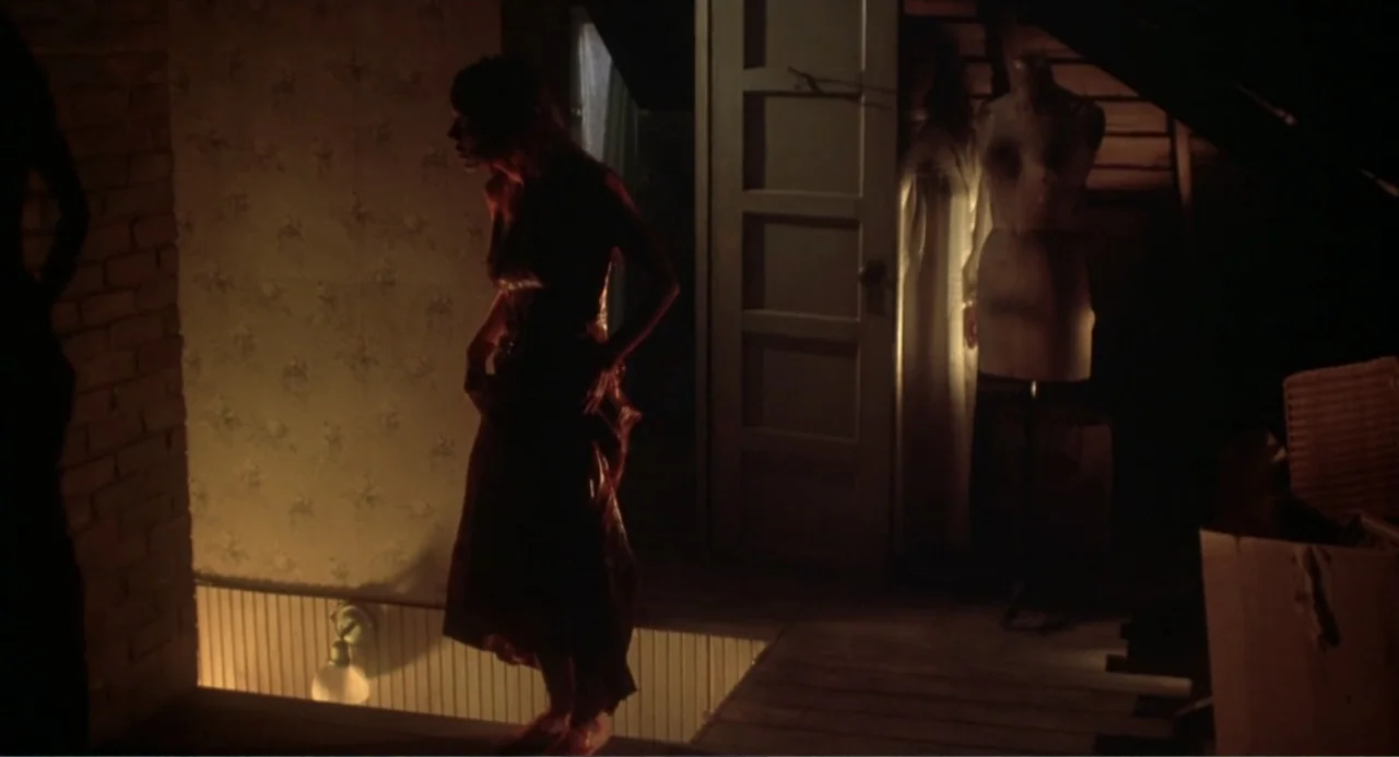 Carrie (1976) movie still: fullbody — A cluttered, dimly lit attic features patterned wallpaper and a sewing mannequin in background. A woman in a…