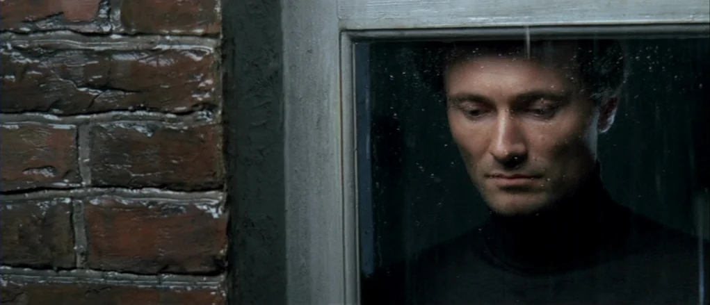 The Banishment (2007) movie still: closeup — A man looks through a pane of glass next to a wet brick wall. Rain streaks are visible on the window…