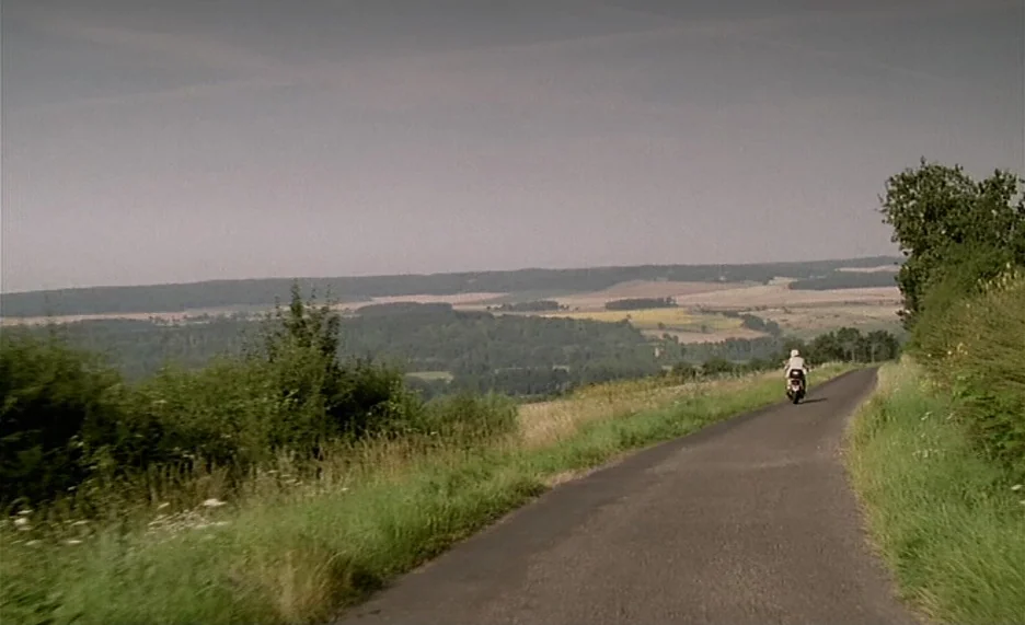Pola X (1999) movie still: wide — An asphalt road winds through a rural landscape with rolling hills and distant trees. A person riding a motorbike…