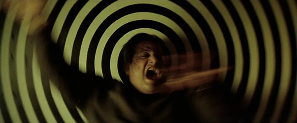 Anguish (1987) movie still: closeup — A man is positioned in front of a high-contrast black and white spiral graphic. The subject gestures wildly…