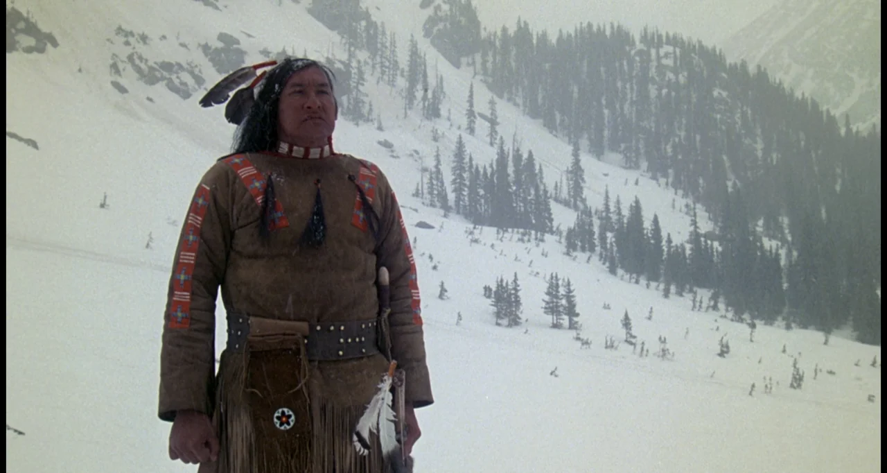 The White Buffalo (1977) movie still: medium — A man stands on a steep, snow-covered mountain slope with coniferous trees nearby. The subject looks…