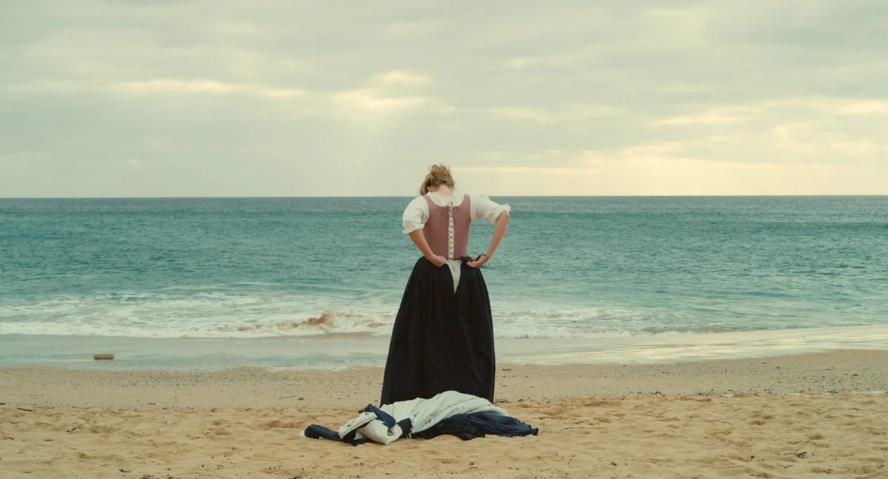 Portrait of a Lady on Fire movie still: fullbody — A sandy coastline under a cloudy sky overlooks a calm sea at daytime. A woman in period clothing…