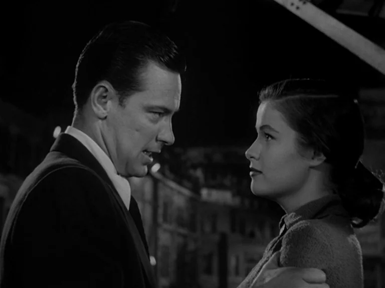 Sunset Boulevard (1950) movie still: medium — A nighttime city street setting with indistinct architectural shapes in the background. A man stands…