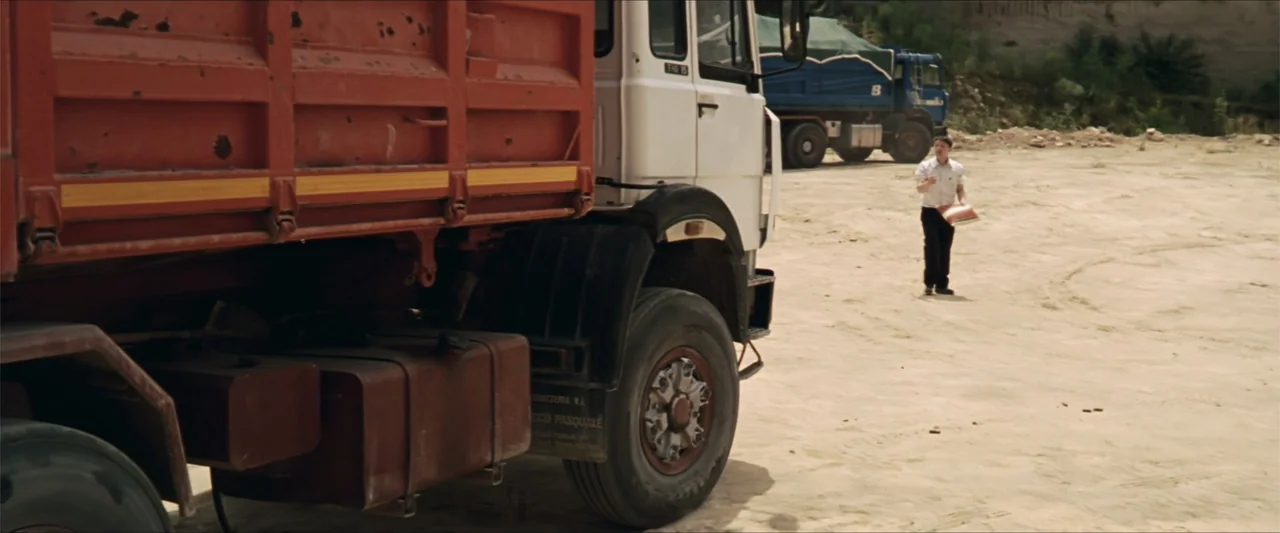 GOMORRAH (2023) movie still: wide — A vast, dusty quarry floor is occupied by heavy trucks and industrial equipment. A man in formal attire stands…