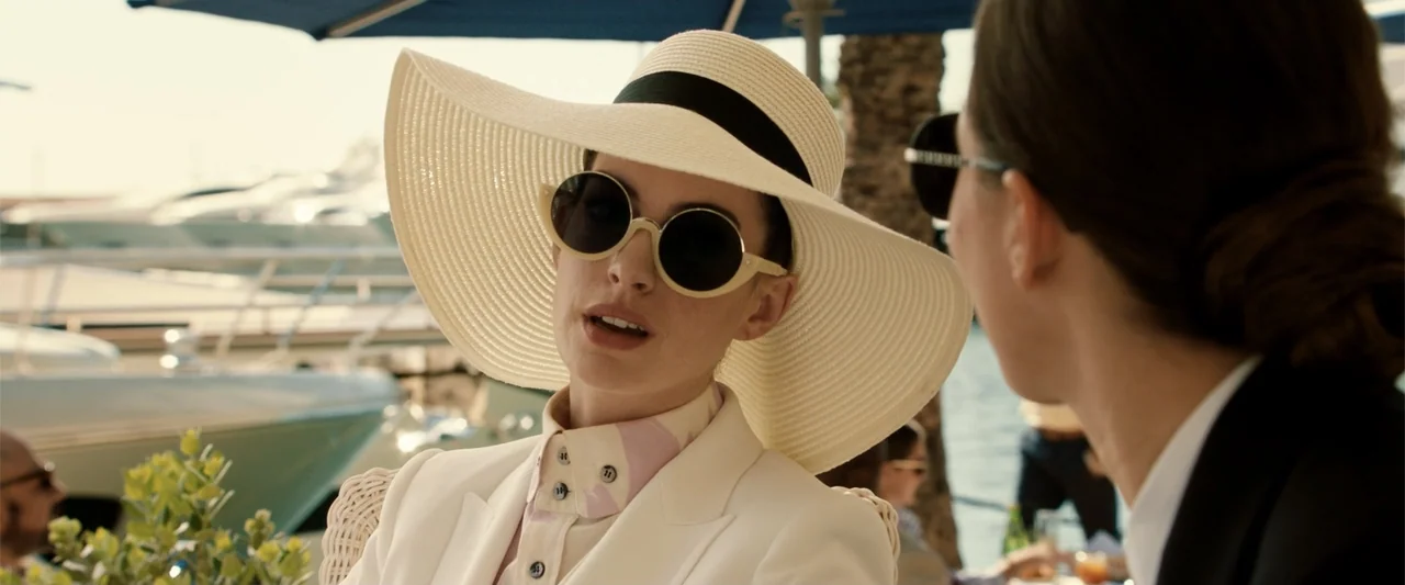 The Hustle (2019) movie still: medium — A sunny marina waterfront with yachts visible in the out-of-focus background. A woman in a large hat talks…